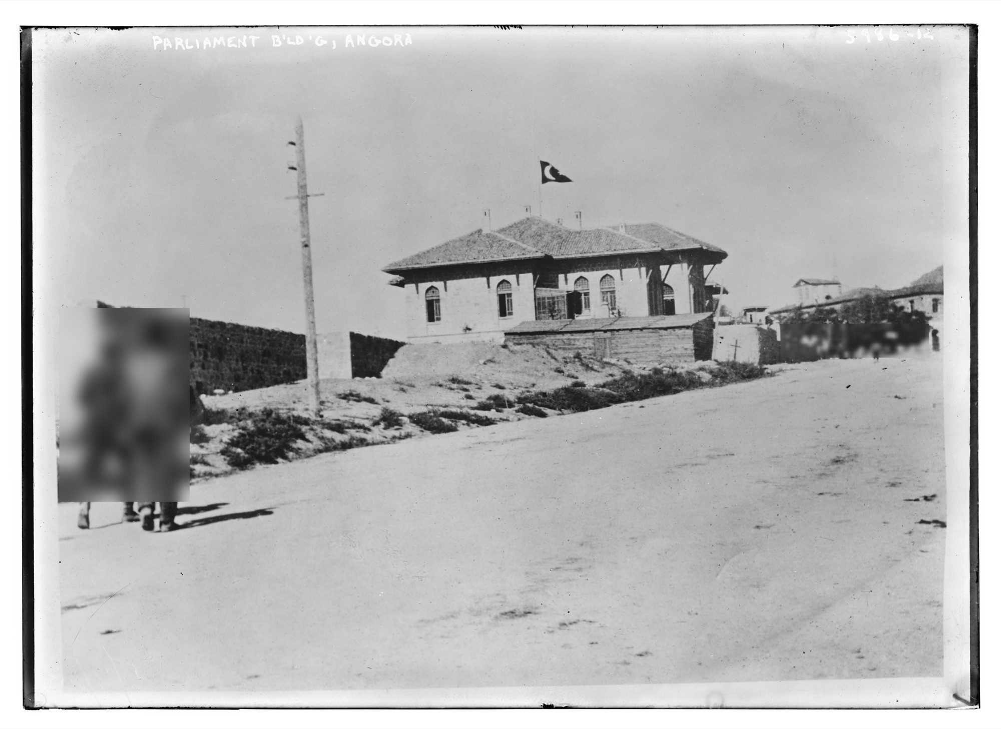 The Bain collection glass negative. A tiled-roof building with pointed-arch windows at the end of a dirt road, a flag on the roof. Two walkers at front left are blurred; their shadows fall on the road.